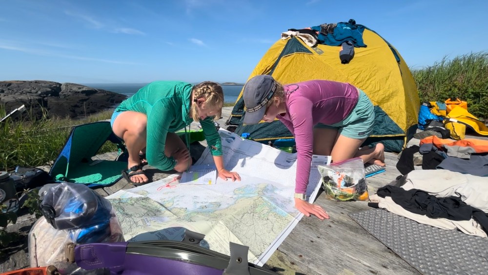 Two campers study a map on a wooden platform, with a yellow tent and scattered gear nearby.