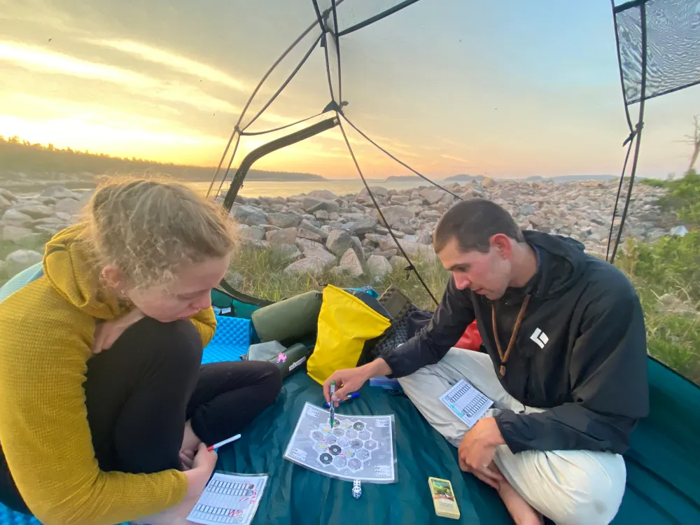 Two people playing a board game in a tent by a rocky shoreline at sunset.