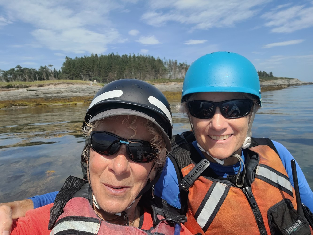 Two people in helmets and vests smile at the camera with a forested shore in the background.