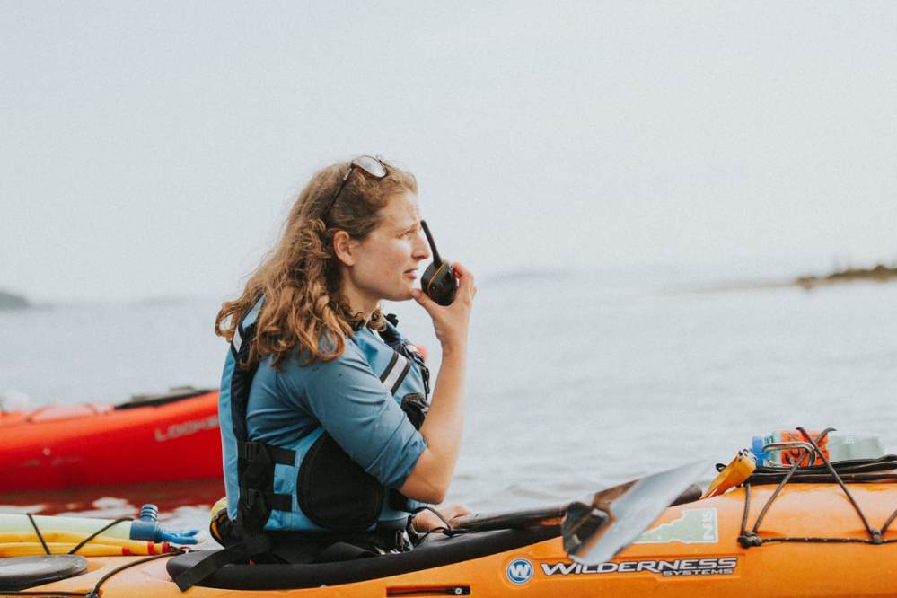 Person in a blue shirt on a kayak using a walkie-talkie on a calm water body.