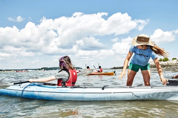 Woman helping person in kayak on sunny day, other kayakers in background.
