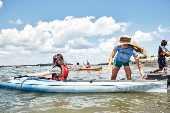People with kayaks on a sunny day near the shore, one wearing a hat helping another. Clouds and other kayaks in background.