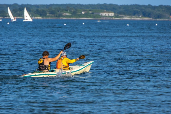Two people kayaking on calm blue water, distant sailboats in background.
