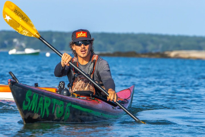 Person kayaking on a calm lake under a blue sky, wearing a cap and sunglasses.