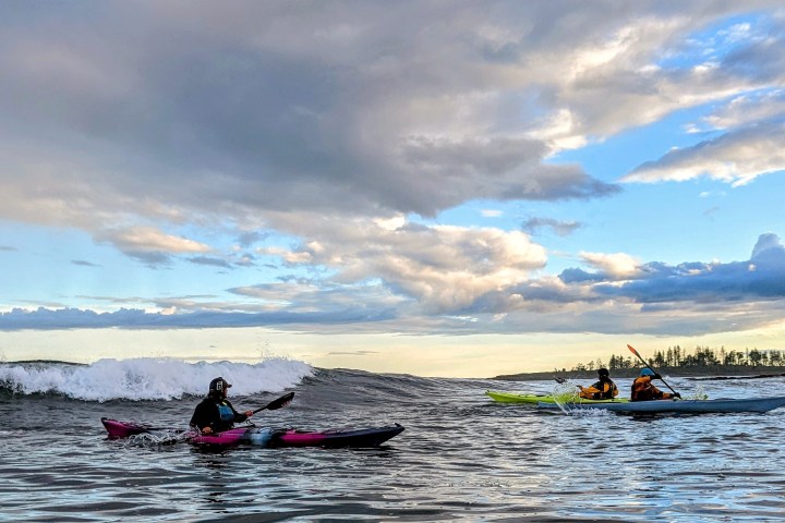 Three kayakers paddling through ocean waves under a cloudy sky.